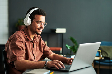 young smiling man in headphones typing on laptop keyboard while sitting by workplace and taking part