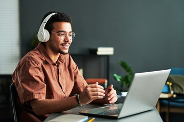 Poster - Young confident student in headphones communicating with online audience or teacher while sitting by desk in front of laptop