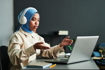 Poster - Young confident Muslim woman in headphones over blue hijab explaining something during online communication