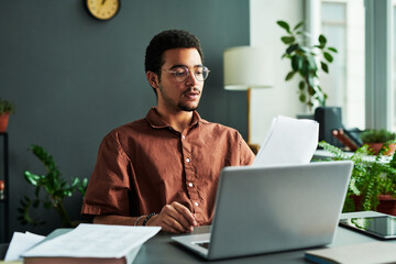 Poster - Young confident man with papers sitting by workplace in front of laptop and making report or explaining new information to online audience