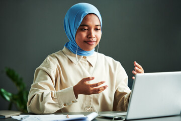 Poster - Young confident Muslim woman in casualwear and blue hijab explaining something while sitting in front of laptop during online lesson
