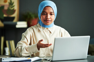 Poster - Young confident Muslim woman in hijab taking part in webinar while sitting by desk in front of laptop at home and explaining something