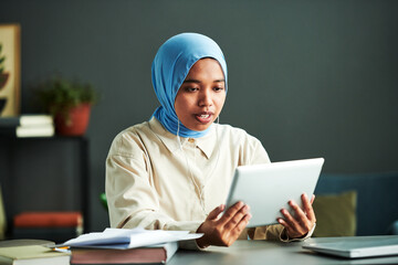Poster - Young confident Muslim female tutor in earphones communicating with online audience while looking at screen of tablet during lesson