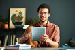 © pressmaster - Young Middle Eastern male student looking through online information on screen of tablet while sitting by workplace with books