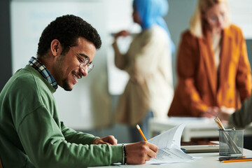 Poster - Side view of happy Middle Eastern student putting ticks in front of right answers while carrying out grammar test by desk in classroom