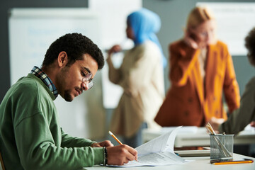 Poster - Young serious student in eyeglasses and casual pullover making notes on paper with exam text during individual work at lesson