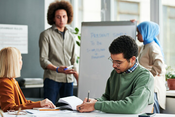 Poster - Young man making notes in copybook while sitting by desk at lesson of English grammar against guy explaining new subject to women