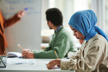 Poster - Side view of young serious Muslim woman in blue hijab making notes in copybook while sitting by workplace during presentation