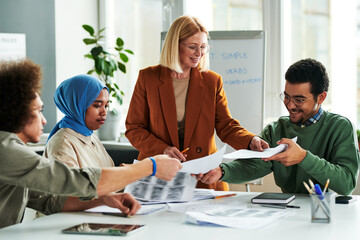 Poster - Happy mature teacher passing papers with grammar tests to group of multicultural students sitting by table at lesson of English language