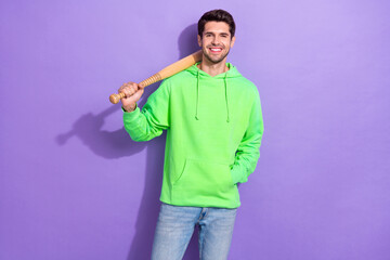 Photo of positive dangerous guy with brunet hairdo dressed green hoodie hold baseball bat on shoulder isolated on purple color background