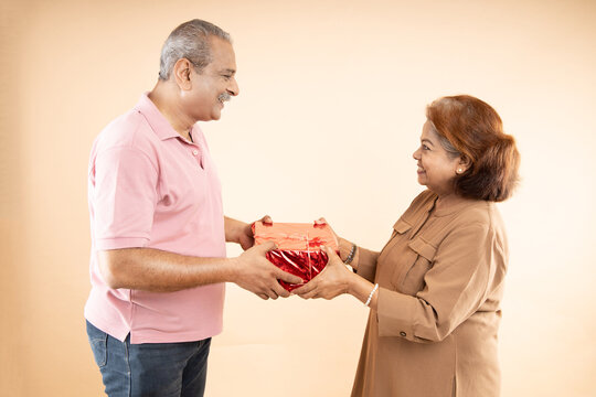 Happy indian senior couple holding gift boxes and celebrating valentine'day together isolated on beige background. Romantic husband give present to his wife to express his love.