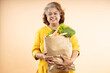 © GAJENDRRA BHATI  - Happy senior indian woman hold grocery paper bags with food isolated on beige background. studio shot.