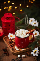 Red cup of hot cocoa with marshmallows on wooden table with christmas tree and glowing garland for christmas