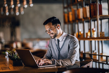 a happy young trendy businessman is typing on a laptop while working remotely from cafeteria.