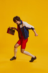 Portrait of man with moustache posing in vintage sportswear and red cycling shorts isolated over yellow background. Dancing with vintage music player