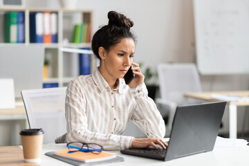 Poster - Serious businesswoman talking on smartphone and typing on laptop keyboard, sitting at workplace in office interior