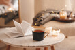 © morrowlight - Cup of tea with paper open book and burning scented candles on marble table over cozy chair and glowing lights in bedroom closeup. Winter holiday season.