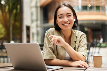 Wall Mural - Urban people. Young woman working with laptop from cafe, coworking space, sitting outdoors with cup of coffee and smiling at camera, holding glasses