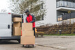 © PoppyPix - Full-length outdoor shot of African-American delivery man using hand truck to deliver cardboard boxes. White delivery van with opened trunk full of packages. Modern building block in the background