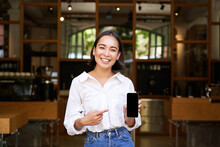 Happy Asian Woman, Cafe Manager Standing In Front Of Restaurant, Pointing Finger At Smartphone App, Showing Mobile Screen, Recommending Application