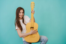 Cheerful Young Girl Laughs, Plays A Yellow Guitar On A Turquoise Background In The Studio, Dressed In A White T-shirt And Blue Jeans, Rocknroll Mood. Mockup, Party Time, Celebrating, Happy People.
