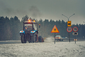 Wall Mural - Tractor with snow plow plowing highway, clearin snow from roadway, prevent slipping on road. Plow tractor plowing snowy roadway. Snow removal, dangerous driving conditions, speed limit sign, icing