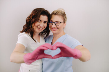 Portrait of two qualified cosmetologists-women, confidently smiling and making a heart with their hands in a medical center