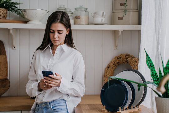 Sad Brunette Caucasian woman in casual standing at kitchen holds phone looks at screen with upset face expression reads message. Female in troubles. Hispanic girl feels loneliness. Failure, divorce.