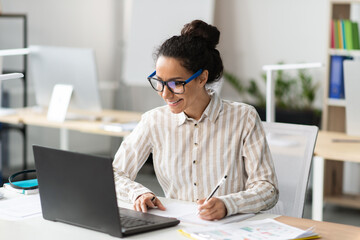 Poster - Happy young latin businesswoman working in office, sitting at workplace, using modern computer and taking notes