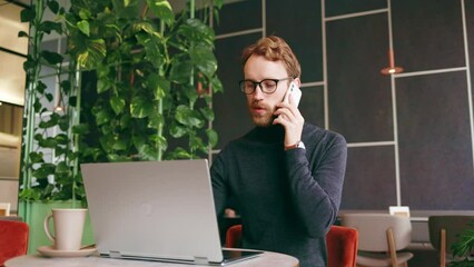 A young red-haired guy, a programmer or entrepreneur in glasses, sits in a stylish cafe at a laptop and speaks on the phone. Freelancer works remotely. Online communication. Small business.