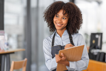 Wall Mural - Female African coffee shop small business owner wearing apron standing in front of counter performing stock check. afro hair employee Barista entrepreneur.