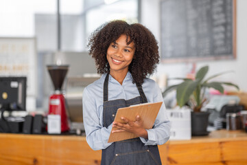 Wall Mural - Female African coffee shop small business owner wearing apron standing in front of counter performing stock check. afro hair employee Barista entrepreneur.