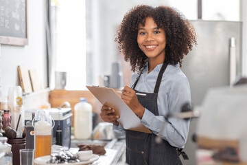 Wall Mural - Female African coffee shop small business owner wearing apron standing in front of counter performing stock check. afro hair employee Barista entrepreneur.