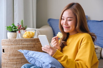 Wall Mural - Portrait image of a beautiful young woman using mobile phone while eating potato chips at home