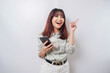 Excited Asian woman wearing sage green suit pointing at the copy space on top of her while holding her phone, isolated by white background