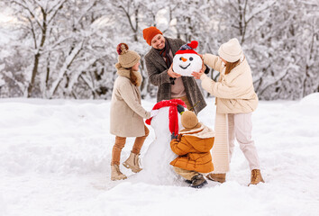 Canvas Print - Happy children sculpting funny snowman together with parents in winter snow-covered park