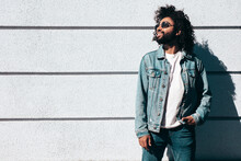 Handsome Smiling Hipster  Model.Sexy Unshaven Arabian Man Dressed In Summer Jeans Jacket Clothes. Fashion Male With Long Curly Hairstyle Posing In Street At Sunset. Cheerful And Happy. In Sunglasses