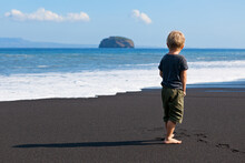 Little Kid Stand On Black Sand Sea Beach. Dreaming Child Look At Sea Surf, Waves. Solitude Concept. Retreat Leisure On Summer Family Vacation