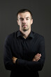 © mikeosphoto - Portrait of man in a black shirt on a dark background. Serious brunet with arms folded. Young caucasian man