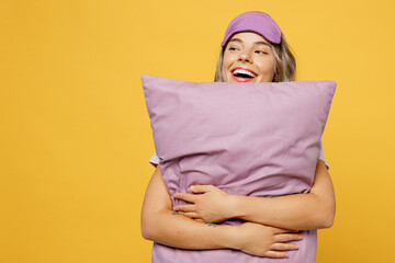 Wall Mural - Fun young woman she wears purple pyjamas jam sleep eye mask rest relax at home hold pillow look aside on workspace area isolated on plain yellow background studio portrait Good mood night nap concept.