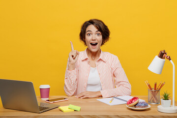 Young employee business woman wear casual shirt sit work at office desk with pc laptop hold index finger up with great new idea isolated on plain yellow color background. Achievement career concept.