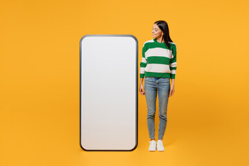 Wall Mural - Full body smiling fun young latin woman wear casual cozy green knitted sweater look at big huge blank screen mobile cell phone smartphone with area isolated on plain yellow background studio portrait.