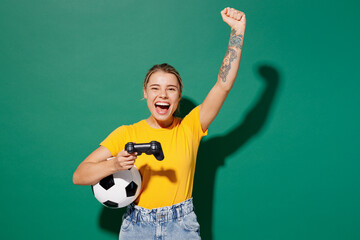 Young winner woman fan in basic yellow t-shirt cheer up support football sport team hold in hand soccer ball watch tv live stream play pc game with joystick console isolated on dark green background