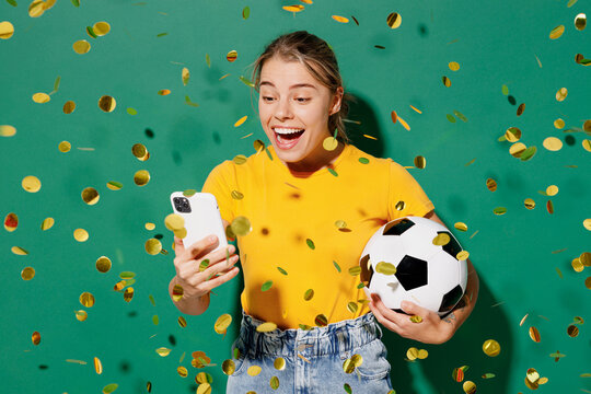 Young woman fan wear yellow t-shirt cheer up support football sport team hold in hand soccer ball watch tv live stream use mobile cell phone among confetti rainfall isolated on dark green background.