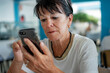 © luciano - Attractive elderly caucasian woman brunette sitting at restaurant table looking on mobile phone the online menu to select food and drink