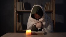Woman Dressed In A Warm Winter Jacket Sit At Home At A Table And Warms His Hands From A Burning Candle. No Heating And Electricity In Winter At Home.