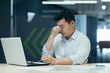 © Liubomir - Tired young Asian businessman. Holds his head, feels fatigue and tension in his head and body, has taken off and is holding his glasses. He sits in the office at a desk with a laptop, needs rest.
