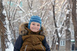 © unona art - Portrait of an attractive boy outdoors in winter. He is wearing a warm hat and jacket, looking at the camera, and smiling. Against the background of trees in the snow and a multi-story building.