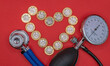 A stethoscope and a sphygmomanometer blood pressure gauge sit on a red background with a heart shape made out of pound coins. Cost of healthcare and the rising expenditure on medical services. 