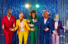 Group Of People In Funny Rubber Masks Of Heads Of Different Animals Are Fooling Around Dancing At Disco Party On Shiny Background. People In Colorful Clothes Wave Hands Funny While Standing In Row.